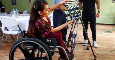 women in wheelchair holding a clapperboard next to camera, two men standing in background