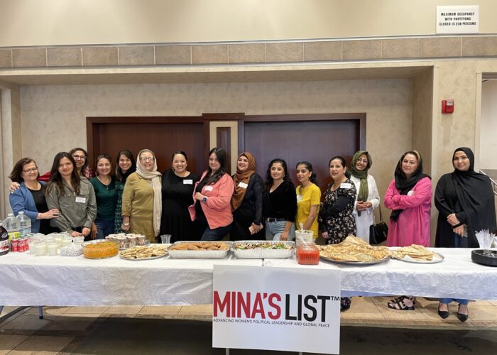 group of women standing in a line behind table with food and drinks and poster that states "Mina's List"