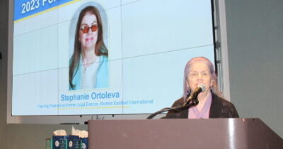 Stephanie Ortoleva standing at a podium, to the right of a large screen presenting her photo and stating "2023 Perdita Huston Honoree".
